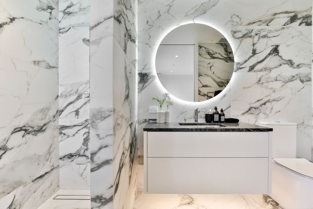 Luxurious contemporary bathroom featuring dramatic white marble-effect wall and floor surfaces with bold grey veining. A floating white vanity with a dark stone benchtop sits beneath a large round backlit mirror, with a wall-hung basin and minimalist black tapware and accessories. The frameless glass shower and partial freestanding bath are visible, creating a bright, refined, high-contrast space.