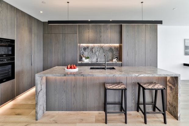 Contemporary kitchen featuring a large waterfall island clad in veined stone, dark timber-grain cabinetry, integrated black appliances, a marble-look splashback, and two woven bar stools. Warm timber flooring and under-cabinet lighting add contrast to the neutral grey and wood palette.