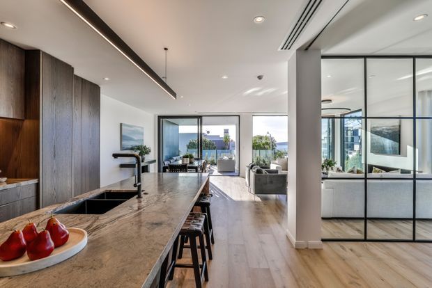 Contemporary open-plan kitchen and living area with a large stone island, dark timber cabinetry, timber flooring, and black-framed glass partitions. The bright interior opens toward a living room and views beyond, with integrated lighting and refined modern detailing.