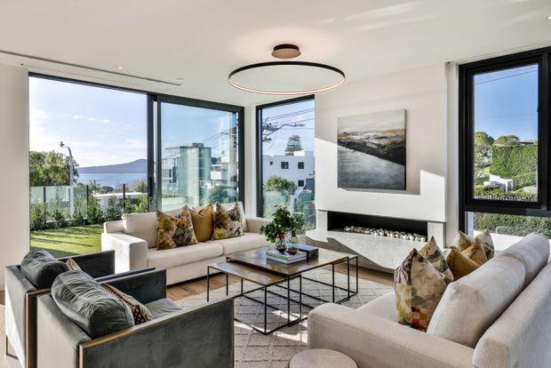 Bright contemporary living room with expansive corner glazing, white upholstered sofas, dark-framed windows, a low black coffee table, built-in fireplace, abstract artwork, and views to a landscaped exterior and distant hills.