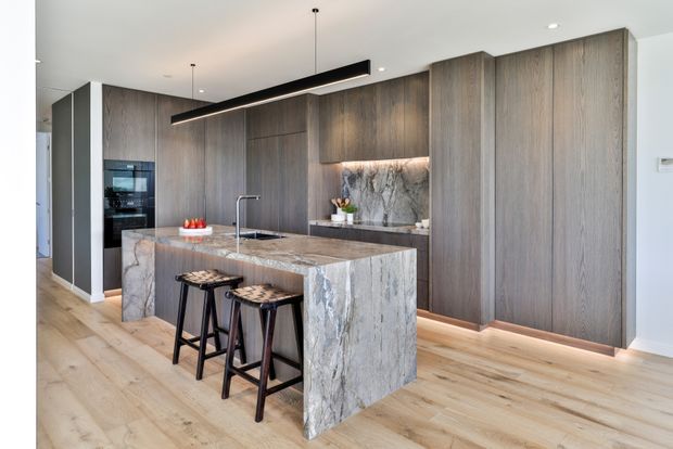A refined contemporary kitchen with a large waterfall-edge stone island, integrated dark timber cabinetry, a stone splashback, timber flooring, and two stools. Recessed lighting and a linear pendant provide layered illumination, while the open layout and concealed appliances create a clean, streamlined appearance.