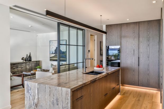 Contemporary open-plan kitchen with a large waterfall-edge stone island, integrated timber cabinetry, black-framed glass partitioning, and concealed warm LED lighting. A living area is visible beyond the partition, while a tall bank of timber joinery and built-in appliances forms the kitchen backdrop.