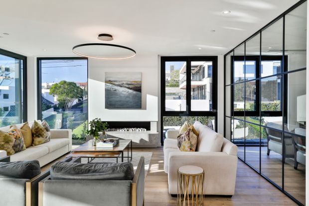 Bright contemporary living room with expansive glazing, black-framed internal glass partitioning, timber flooring, neutral sofas, and a sculptural circular ceiling light. A compact work area is visible behind the glazed partition, with an urban residential outlook through the windows.