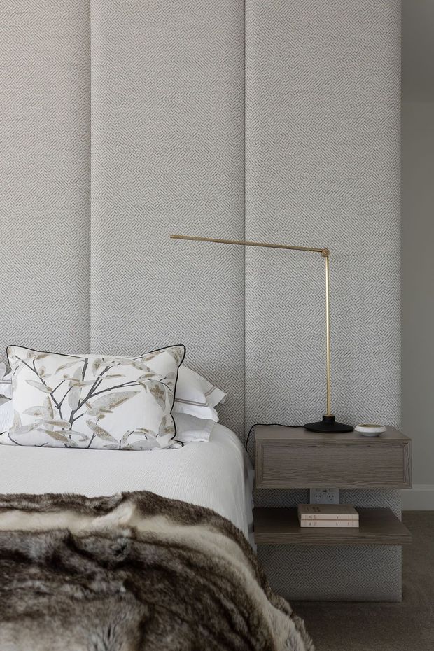 Close-up of a refined bedroom corner featuring a neatly made bed with a botanical-patterned cushion and a dark fur throw, a textured grey upholstered feature wall, and a compact dark wood bedside table with open shelving. A slender brass and black table lamp adds a warm metallic accent. The palette is muted and sophisticated with layered natural textures.