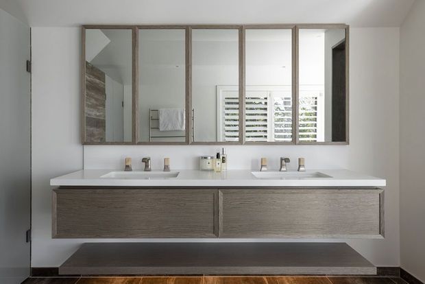 A bright, contemporary double-vanity bathroom with a long white countertop, two integrated rectangular basins, wall-mounted timber-look vanity drawers, and mirrored cabinet doors above. A shuttered window and tiled shower area are visible in the reflections. The palette is predominantly white and light grey with warm wood-grain joinery and dark flooring.