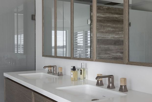A contemporary double-basin bathroom vanity with a white solid-surface countertop, integrated rectangular sinks, brushed metal faucets, and dark wood-grain cabinetry. A large framed mirrored wall cabinet spans the vanity, with a textured wood-toned central panel and bright natural light filtering through shutters.