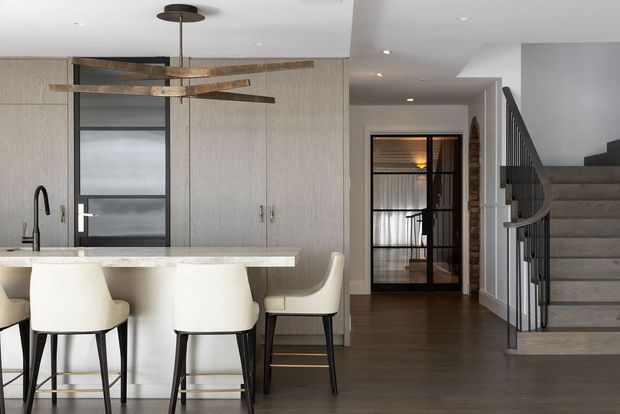 A contemporary open-plan kitchen and circulation area featuring a long pale stone island with white upholstered bar stools, integrated light wood cabinetry, a black-framed glazed door, dark timber flooring, and a sculptural suspended pendant light. A stairway with dark metal balustrading rises on the right, while a glazed internal door opens toward a hallway or adjoining room.