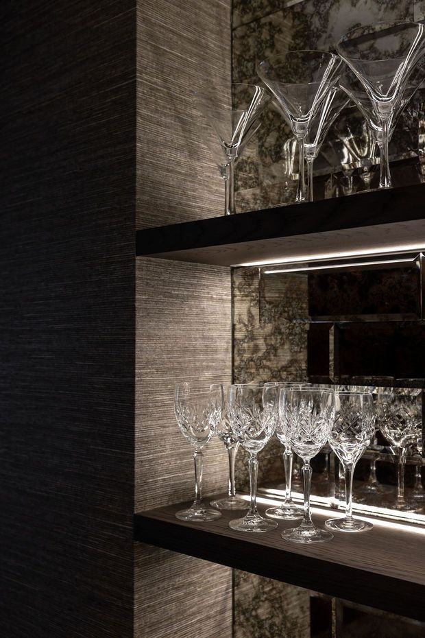Close-up of a sophisticated built-in glassware display cabinet or bar storage niche. Crystal wine glasses and martini glasses are arranged on dark shelves, with mirrored backing and integrated linear lighting. The surrounding wall finish has a dark, horizontally textured appearance, creating a moody and luxurious atmosphere.