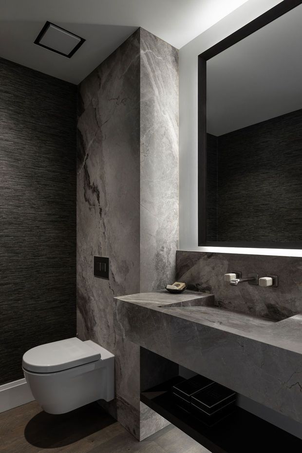 A compact, dramatic contemporary bathroom with dark textured wall finishes, large grey stone-look slabs, a wall-hung toilet, and a floating stone vanity with integrated basin. A large black-framed illuminated mirror and concealed wall-mounted tapware complete the refined, monochromatic design.