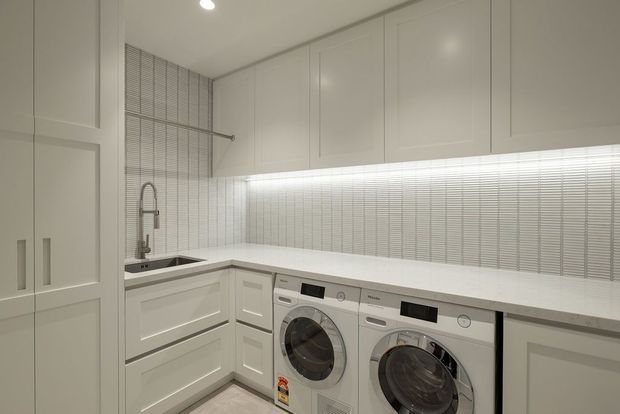 Bright, compact contemporary laundry room with white built-in cabinetry, a white benchtop, integrated front-loading washer and dryer, stainless-steel sink and tap, and a textured horizontal tile splashback. Recessed ceiling lighting and a clean neutral palette create a highly functional, streamlined fit-out.