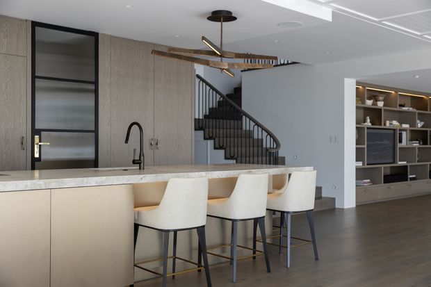 Contemporary open-plan kitchen and living area featuring a large pale stone island with four upholstered bar stools, integrated light cabinetry, a black-framed reeded-glass door, timber-toned wall cabinetry, a black metal staircase, and built-in living-room shelving. A sculptural linear pendant adds a warm brass and dark accent above the island.