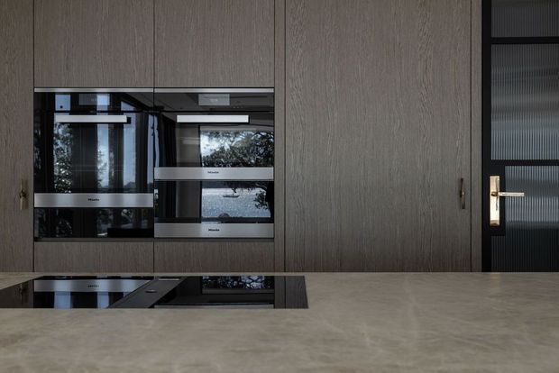 A refined contemporary kitchen featuring full-height dark timber-grain cabinetry, integrated built-in ovens, a black induction cooktop, and a broad stone-look island benchtop. A black-framed ribbed-glass door with a brass lever adds contrast at the right.