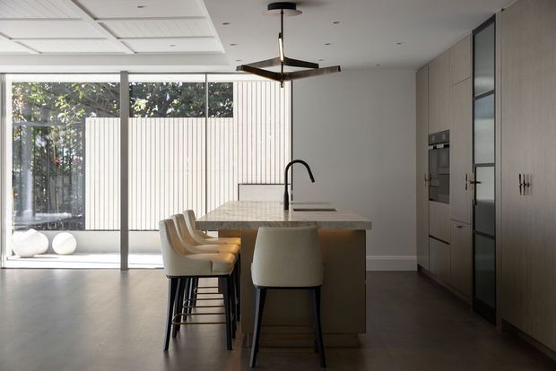 Contemporary open-plan kitchen featuring a large stone island with integrated sink and dark gooseneck tap, upholstered bar stools, full-height light timber cabinetry, and integrated appliances. Floor-to-ceiling glazing opens to a bright courtyard or outdoor area, while a minimalist ceiling fan and recessed lighting complete the space.