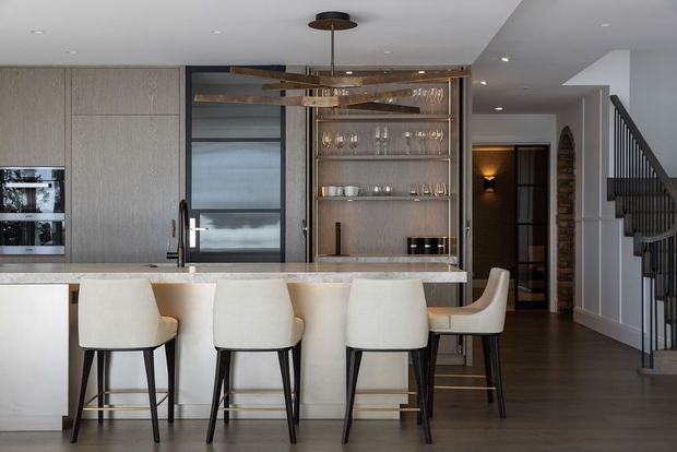 Contemporary open-plan kitchen with a large stone island and four upholstered bar stools, vertically grained cabinetry, integrated appliances, a dark-framed glazed pantry or storage door, and open shelving displaying glassware. A sculptural brass-toned pendant hangs above the island. The space opens toward a hallway and staircase with dark timber flooring and neutral finishes.