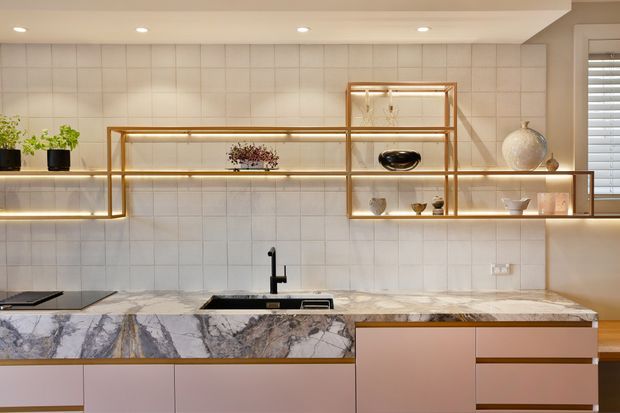 Contemporary kitchen with a full-height pale tiled splashback, dramatic veined stone benchtop, blush-toned flat-panel cabinetry, and open brass-framed shelving with integrated warm lighting. A black sink mixer and cooktop provide contrast, while curated ceramics and greenery style the display shelves.