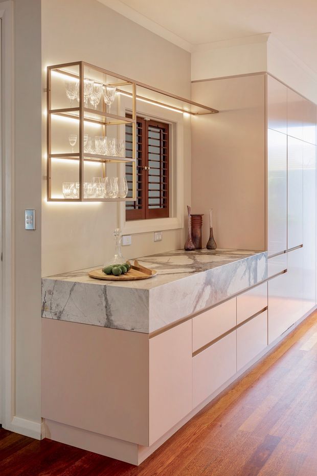 A refined built-in bar or drinks station in a warm-toned interior, featuring pale cabinetry, a dramatic veined stone benchtop, integrated LED lighting, and open bronze-toned shelving displaying glassware. A timber-framed window with security bars sits behind the counter, while polished timber flooring adds warmth.
