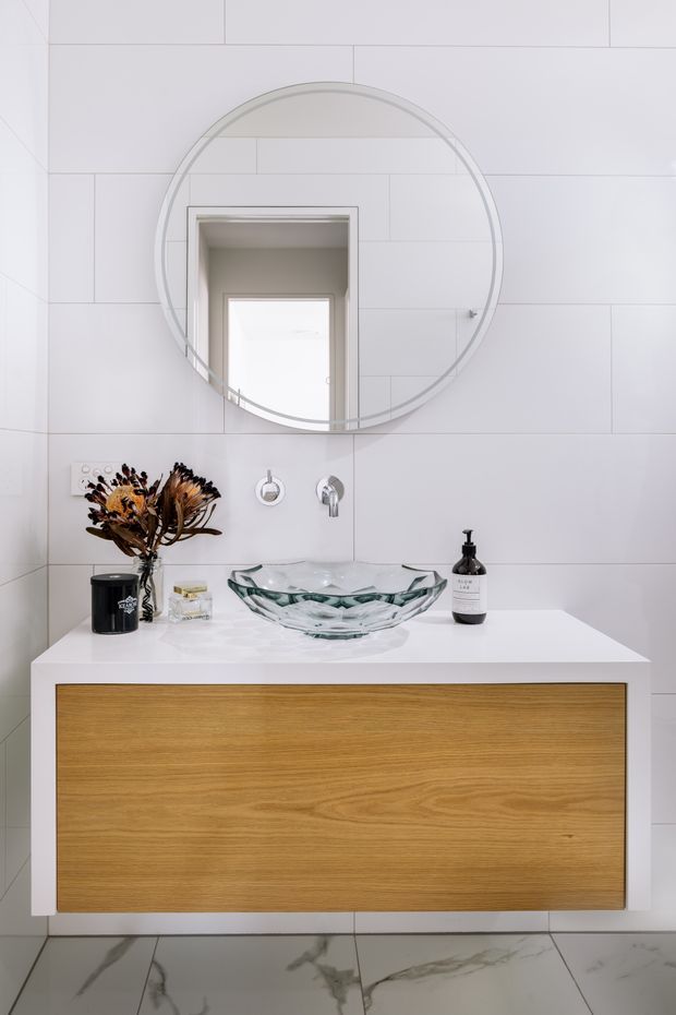Contemporary bathroom vanity with a large round frameless mirror, white rectangular wall tiles, a white countertop, a clear glass vessel basin, wall-mounted chrome tapware, and a floating warm timber-front cabinet. Large marble-look floor tiles and minimal black accessories complete the clean, refined scheme.
