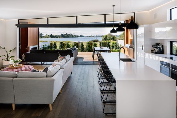 Bright open-plan contemporary interior combining a white kitchen with a large island and bar seating, a furnished living area, and expansive glazing opening to a timber deck with a landscaped waterfront outlook. Dark timber flooring, white cabinetry, black-framed glazing, and pendant lights create a clean, refined scheme.