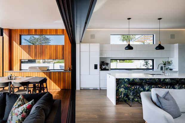 Open-plan contemporary kitchen, dining, and living area with warm vertical timber wall panelling, white handleless cabinetry, a large island with a dark botanical-patterned front, timber flooring, black pendant lights, and extensive internal glazing or sliding doors. A dining table and sofas are visible, with bright natural light throughout.
