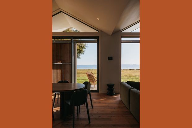 A warm, compact contemporary interior combining a dining area and living space, with dark timber flooring, a round dining table and upholstered chairs in the foreground, and a sofa to the right. Full-height glazed doors and high clerestory-style glazing open the room to a rural coastal landscape and broad water views. The interior has warm neutral walls, dark framed glazing, timber screening, and a calm, refined atmosphere.