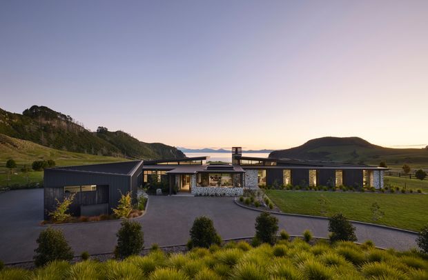 Wide exterior view of a low-slung contemporary residence set in a landscaped rural or mountainous site at dusk. The home features dark cladding, extensive glazing, a long horizontal roofline, a central stone-clad entry volume, and a separate garage or outbuilding connected by a paved driveway. Rolling hills and distant mountains form the background.
