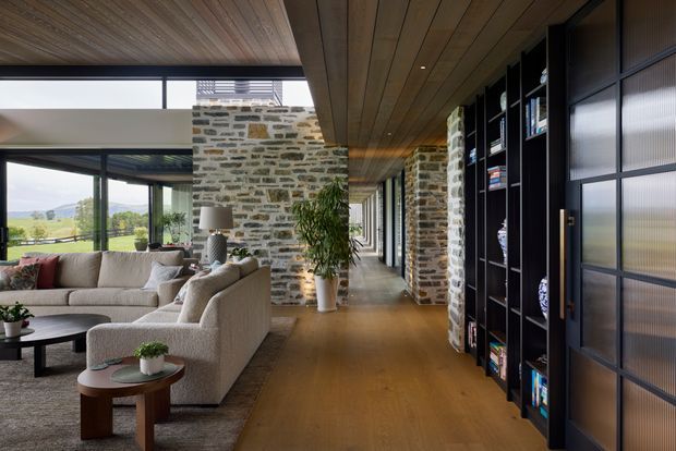 A refined open-plan living room and circulation corridor with exposed rough stone walls, warm timber floors and a timber-lined ceiling. A large neutral sectional sofa sits beside expansive glazing overlooking a green landscape, while a long wall of dark built-in shelving and translucent-panel doors runs along the hallway. The space combines natural materials, strong horizontal lines and abundant daylight in a contemporary rural setting.