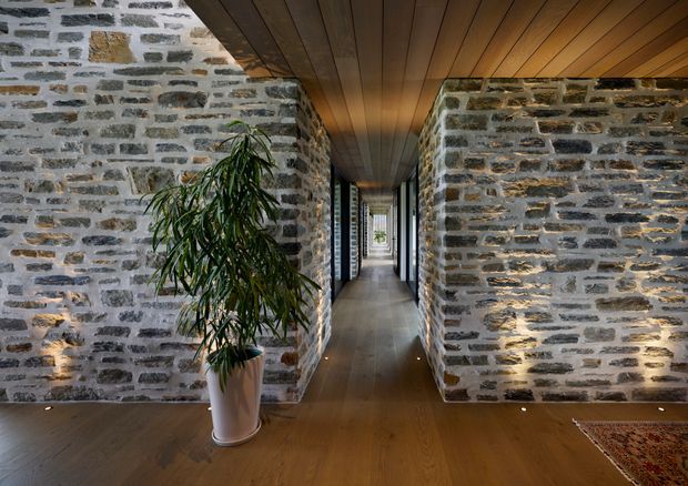 A dramatic interior hallway with dark timber flooring and a matching timber-lined ceiling, flanked by exposed grey stone masonry walls. Recessed uplighting washes the stone surfaces, while a tall potted plant and a glimpse of a patterned rug add softness. The corridor extends toward a bright glazed opening at the far end.
