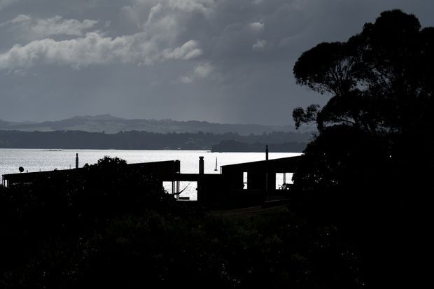 A dark, high-contrast silhouette of a low waterfront building or pavilion viewed from shore, with a broad body of water, distant rolling hills, dramatic clouds, dock-like vertical posts, and a large tree framing the right side. Architectural details are mostly obscured by backlighting.