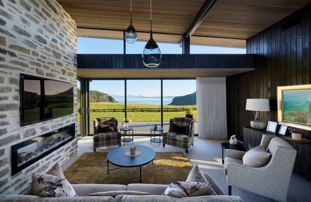 A refined double-height living room with expansive floor-to-ceiling glazing overlooking a rural valley and distant mountains. The room features a dramatic rough-stone fireplace wall with integrated television and shelving, dark vertical timber wall panelling, a warm timber-lined ceiling, exposed dark structural beams, pendant lights, a large sectional sofa, armchairs, a round coffee table, and a mezzanine-like upper-level walkway. The interior blends natural stone, timber, dark finishes, and soft neutral furnishings in a warm contemporary setting.