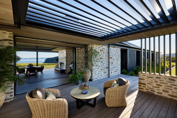 A covered outdoor living and dining area with a contemporary slatted pergola roof, timber decking, exposed pale stone walls, and vertical glazed openings. Wicker lounge chairs and a round coffee table create a relaxed seating zone, while a dining table sits beyond in an open-air section. The space overlooks a coastal landscape with water and headland views, and includes lush potted planting and integrated architectural glazing.