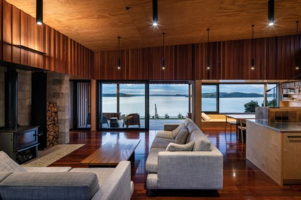 A warm, richly detailed open-plan living room overlooking a body of water through extensive floor-to-ceiling glazing. The room features dark polished timber flooring, timber-clad ceiling and walls with vertical slatted detailing, a large sectional sofa, a wood-burning stove with stacked firewood, and an integrated timber kitchen or bar joinery area. The interior has a refined contemporary lodge-like character with strong indoor-outdoor visual connection.