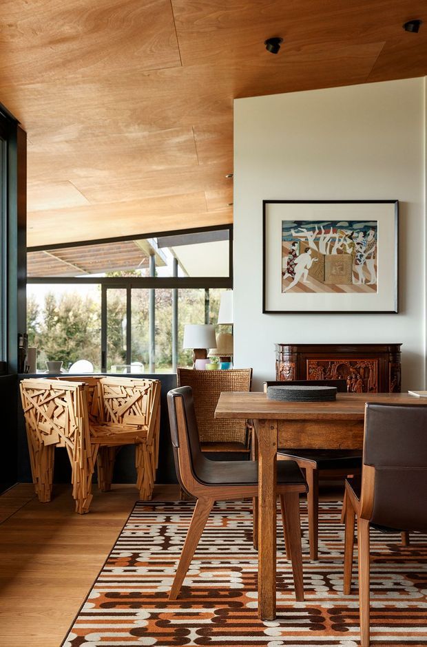 A warm, richly detailed dining area with a substantial timber dining table and sculptural timber-and-leather chairs. The room features a wood-clad ceiling, dark-framed floor-to-ceiling glazing, a patterned area rug, a carved timber sideboard, framed artwork, and woven occasional furniture. Natural light enters through the glazing, with an outdoor covered area visible beyond.