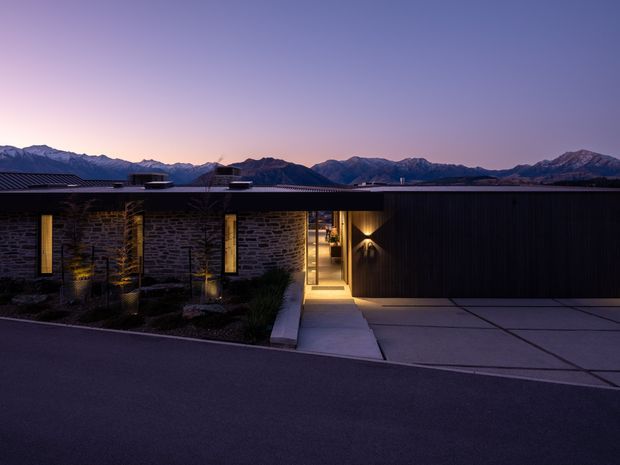 Twilight exterior view of a low, modern single-storey home set against a prominent mountain range. The facade combines dark horizontal cladding with irregular natural stone masonry, narrow vertical windows, warm recessed lighting, and a sheltered entry walkway. A broad concrete driveway occupies the foreground, while the flat or very low-pitched roofline and restrained landscaping reinforce the contemporary architectural character.