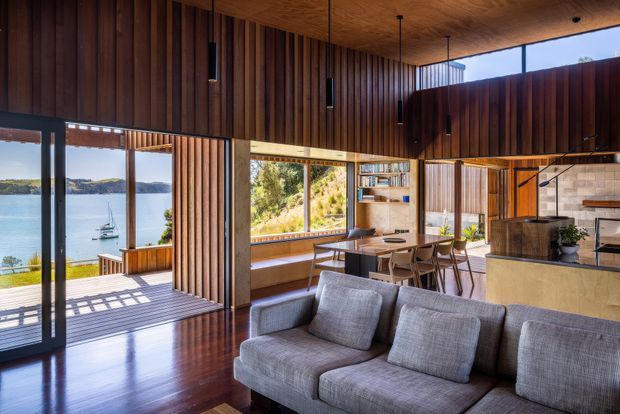 A warm, contemporary open-plan living space with a large grey sectional sofa in the foreground, a timber dining area and kitchen beyond, and extensive glazing opening to a timber deck. Vertical timber wall panelling and a timber ceiling create a strong natural-material interior, with expansive views over a sheltered body of water and surrounding landscape.