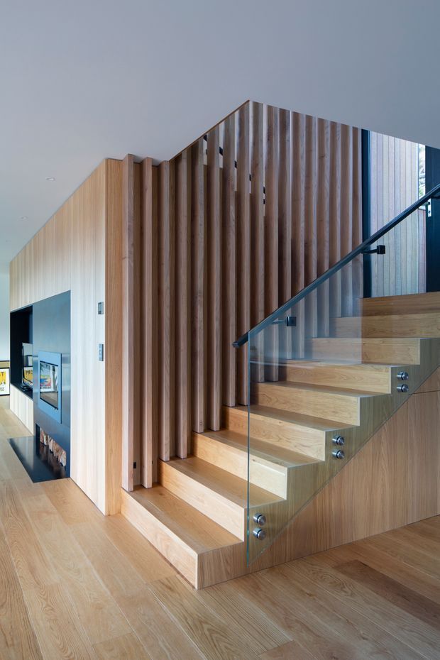 Interior architectural photograph showing a contemporary timber staircase with broad floating wooden treads, clear glass balustrade, and a dark metal handrail. Vertical timber battens form a sculptural screen along the stair, with matching timber wall cladding and flooring. A glimpse of an adjoining living area is visible at left.