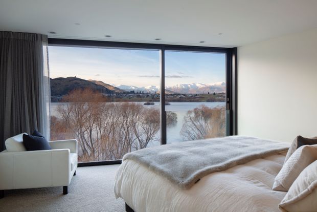 A serene modern bedroom with a large floor-to-ceiling corner window overlooking a lake, wooded shoreline, and snow-capped mountains. The room features a low-profile upholstered bed layered with white bedding and a grey throw, a white armchair with a dark cushion, full-height dark curtains, recessed ceiling lights, and soft neutral finishes.