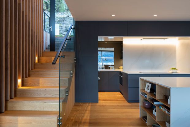 Contemporary open-plan kitchen and stairway interior with warm timber flooring and timber-lined stair treads, a sculptural glass balustrade, dark charcoal cabinetry, and a pale stone island or benchtop. Vertical timber battens line the stair wall, while integrated lighting creates a warm, refined atmosphere. A glimpse of exterior landscape is visible through the glazing.