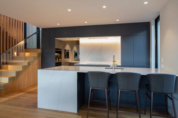 Contemporary open-plan kitchen with a large pale stone island and integrated breakfast bar, dark matte cabinetry, warm timber flooring and a sculptural floating timber staircase with glass balustrade. Recessed ceiling lighting and concealed under-cabinet illumination create a refined, softly layered interior.