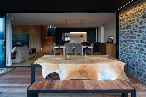 Open-plan interior featuring a dining area and kitchen, with a living room visible to the left. The space combines dark timber cabinetry, black-framed glazing, a rustic exposed stone feature wall, timber flooring, and a substantial dining table with a natural hide covering. A staircase and pendant lighting add architectural definition.