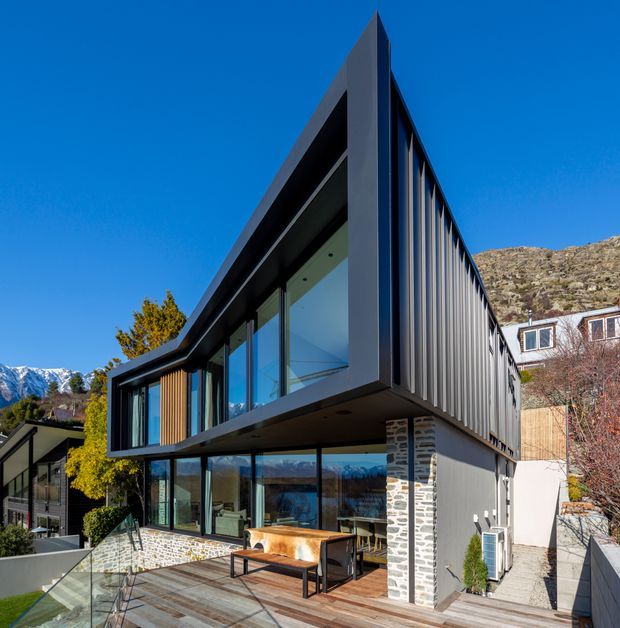 Exterior view of a contemporary multi-level house on a sloping site, featuring a sharply angular projecting upper storey, extensive floor-to-ceiling glazing, dark vertical metal cladding, timber accents, a stone-clad structural column, and an outdoor terrace. Mountainous terrain and neighbouring residences are visible in the background.