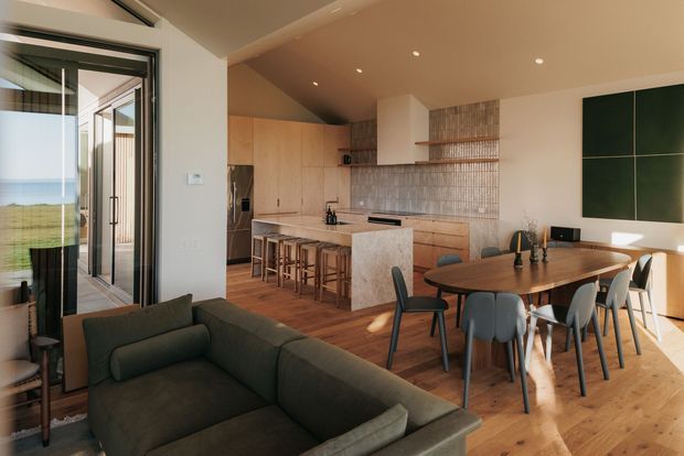Open-plan contemporary kitchen, dining, and living area with warm timber flooring and cabinetry, a large stone island, tiled splashback, timber dining table, and dark upholstered seating. Full-height glazed doors open to a grassy coastal landscape with ocean visible beyond. Recessed ceiling lighting and dark-framed glazing create a refined, modern interior.