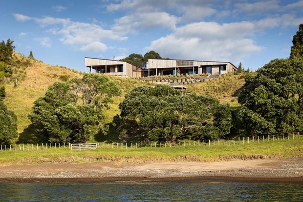 A low-slung contemporary residence positioned on a grassy, elevated coastal or waterfront site. The building is composed of connected rectangular volumes with simple metal-clad walls, broad glazing, and shallow pitched or nearly flat rooflines. Terraced landscaping and a long line of outdoor seating or retaining elements follow the hillside, with trees, pasture fencing, a narrow shoreline, and water in the foreground.