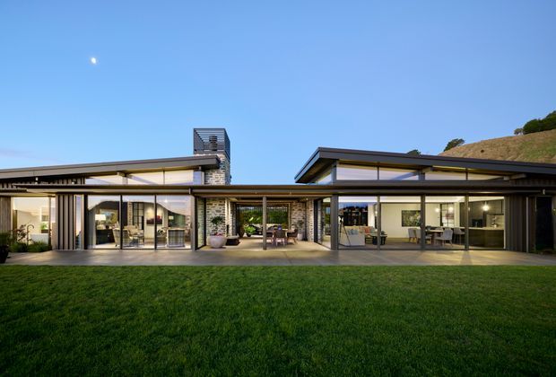 A wide exterior view of a low-profile contemporary residence with extensive floor-to-ceiling glazing, dark slim metal framing, pale masonry walls, and broad shallow roof planes. The home opens onto a covered central courtyard or terrace, with living and dining areas visible through the glass. A manicured lawn occupies the foreground, and the building is set against a sloping landscaped site under a clear blue sky.