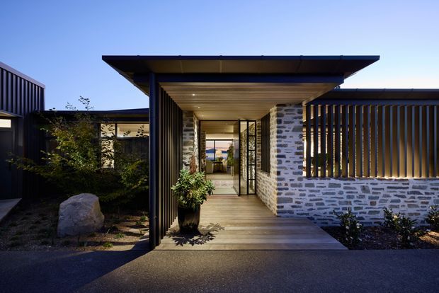 Contemporary residential entry viewed from outside at dusk, featuring a broad flat roof canopy, warm timber soffit and decking, dark vertical slatted screens, and a low stone-clad wall with a glazed doorway leading into the home. Landscaping and a large boulder frame the sheltered walkway.