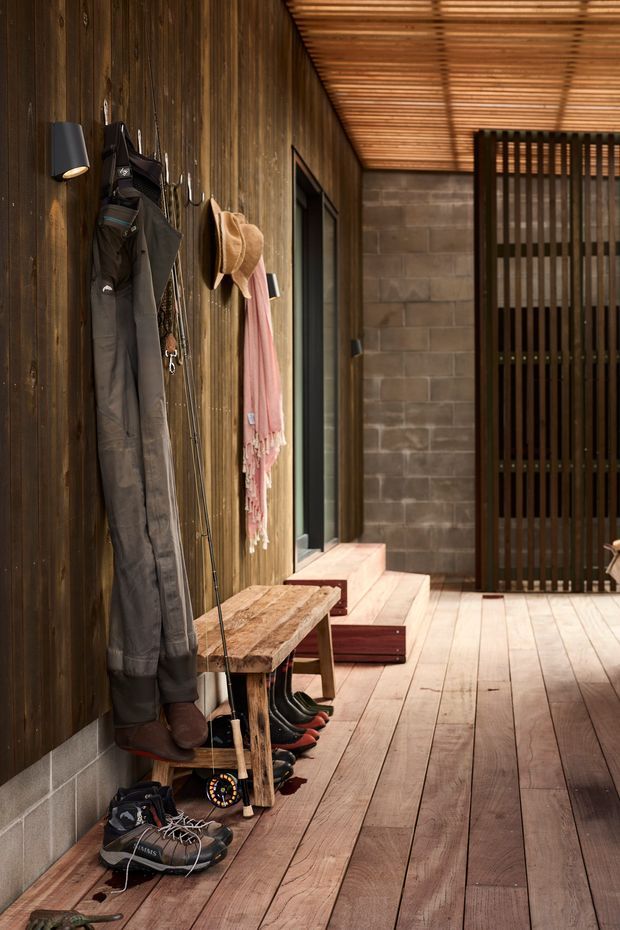 A warm, rustic covered entry corridor with dark vertical timber wall cladding, timber plank flooring and a slatted timber ceiling. A built-in bench, hooks, outdoor clothing, hats, boots and sporting gear line the wall, while a concrete block wall and vertical timber screen define the far end. The space has a practical mudroom-like entry character with strong natural materials and layered timber detailing.