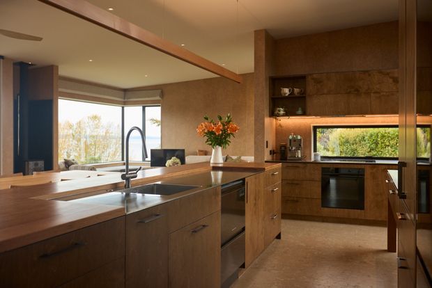 Warm, refined contemporary kitchen with extensive dark timber cabinetry, a large timber island, integrated stainless-steel sink and tapware, built-in oven, and a horizontal garden-facing window. The kitchen opens to a bright living area with large glazing and built-in shelving, creating a spacious open-plan interior.