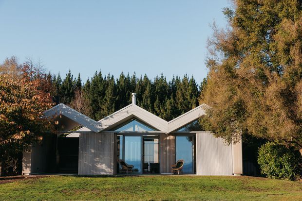 Single-storey contemporary house set within a landscaped, tree-lined site. The facade features a distinctive repeated zigzag pitched roof profile, vertical light-grey cladding, large glazed openings, and a central glazed entry or living area. Outdoor chairs are visible beneath the covered portions of the facade.