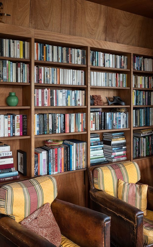 A richly detailed reading area dominated by a full-height, dark timber built-in bookshelf filled with books. Two vintage leather armchairs with patterned mustard, green, and red upholstery sit in front of the shelving, with decorative objects and cushions adding warmth. The space has a cosy, book-lined library atmosphere and traditional crafted joinery.