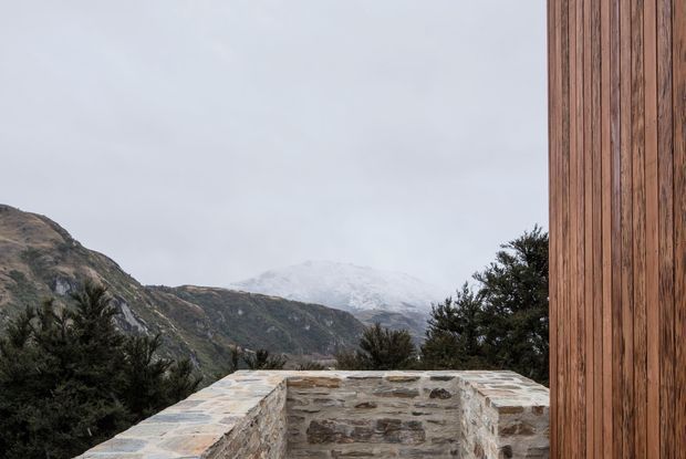 Exterior architectural view featuring a low, rough stone parapet or terrace wall in the foreground, a tall vertical timber-clad facade at right, and rugged mountainous terrain visible beyond under an overcast sky. The composition highlights the building materials and its mountain setting.