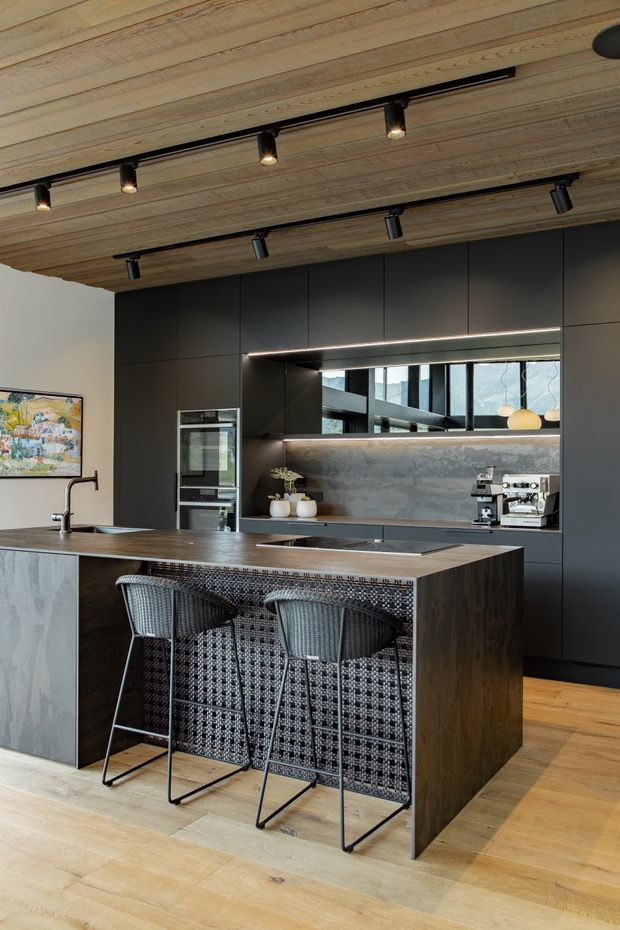 Contemporary kitchen with a large dark island, integrated matte-black cabinetry, stone-look splashback, built-in appliances, and a woven-texture island front with two bar stools. The room features a warm timber-lined ceiling with black track lighting, timber flooring, and a long horizontal window providing natural light.
